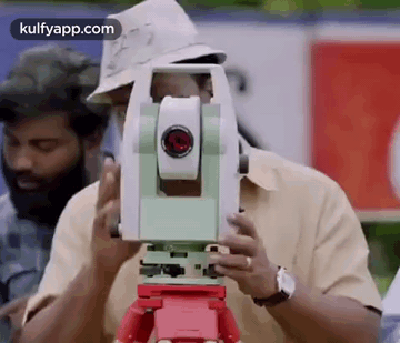 A Man In A Hat Is Using A Theodolite On A Tripod To Measure Something . GIF