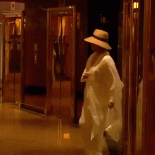 A Man In A Hat Is Walking Down A Hallway Holding A Bag . GIF