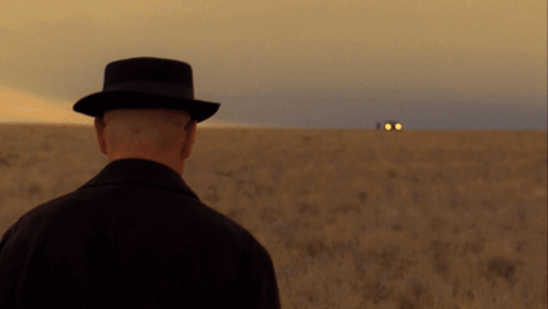 A Man In A Hat Stands In A Field Looking At A Car GIF