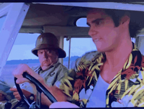 A Man In A Hawaiian Shirt Is Driving A Car With Another Man In A Safari Hat Behind Him GIF