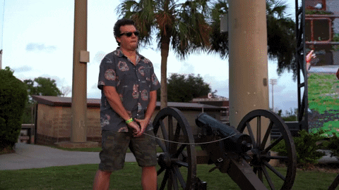 A Man In A Hawaiian Shirt Is Standing Next To A Cannon In A Park GIF