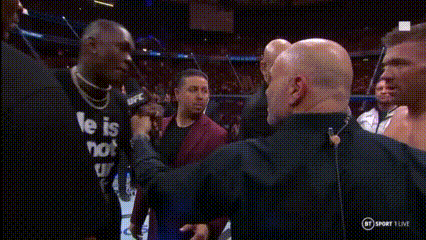 A Man In A He Is Not Shirt Is Talking Into A Microphone While Another Man Looks On . GIF
