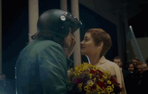 A Man In A Helmet Kisses A Woman Holding Flowers GIF