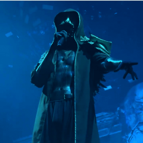 A Man In A Hooded Coat Is Singing Into A Microphone In A Dark Room GIF