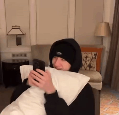 A Man In A Hooded Jacket Is Holding A Pillow And Looking At His Phone . GIF