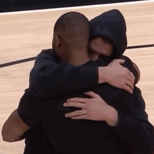 A Man In A Hooded Jacket Is Hugging Another Man On A Basketball Court . GIF