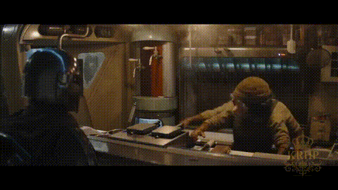 A Man In A Hooded Jacket Is Sitting At A Table In A Dark Room With A Crown On It . GIF