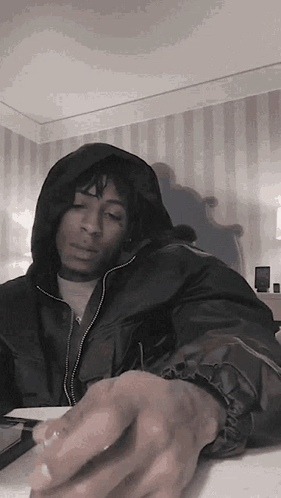 A Man In A Hooded Jacket Is Sitting At A Table With His Hands On The Table . GIF