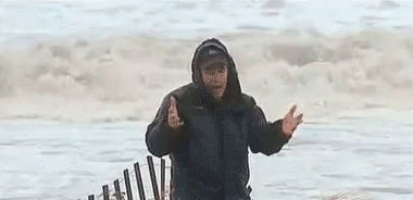 A Man In A Hooded Jacket Is Standing In Front Of A Fence On The Beach . GIF