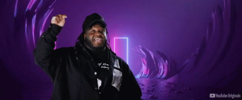 A Man In A Hooded Jacket Stands In Front Of A Sign That Says Mi Escena Musical GIF