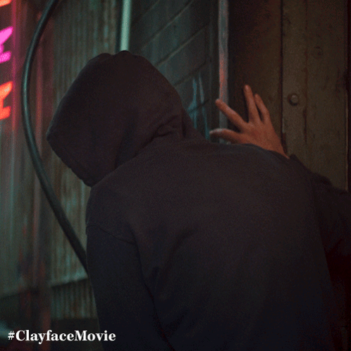 A Man In A Hooded Sweatshirt Is Standing In Front Of A Neon Sign That Says Clayface Movie GIF