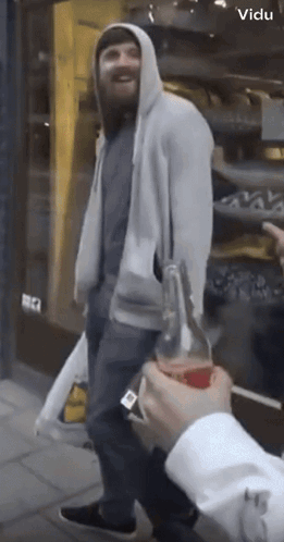 A Man In A Hoodie Holds A Bottle Of Soda In Front Of A Store That Says Vidu On It GIF