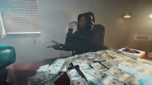 A Man In A Hoodie Is Sitting At A Table With A Pile Of Money On It . GIF