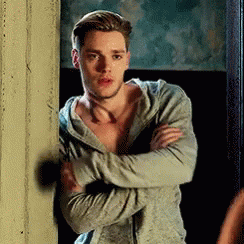 A Man In A Hoodie Is Standing Next To A Wall With His Arms Crossed And Looking At The Camera . GIF