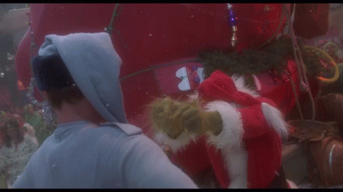 A Man In A Hoodie Talks To A Grinch Who Is Wearing A Santa Hat GIF