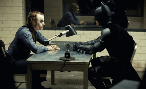 A Man In A Joker Costume Sits At A Table With A Batman Costume GIF