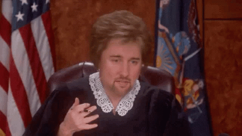 A Man In A Judge 'S Robe Is Sitting In Front Of A Flag . GIF
