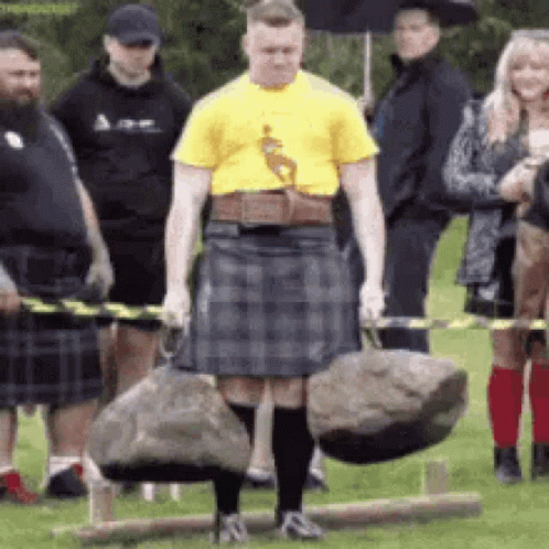 A Man In A Kilt Is Carrying A Large Rock While A Group Of People Watch . GIF