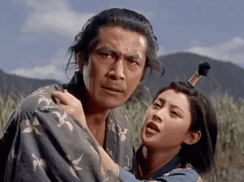 A Man In A Kimono Is Holding A Woman In A Field With Mountains In The Background GIF