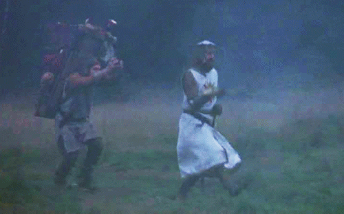 A Man In A Knight 'S Costume Is Walking Through A Field With Another Man Carrying A Backpack . GIF