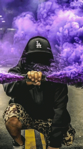A Man In A La Hat Holds A Purple Smoke Bomb GIF