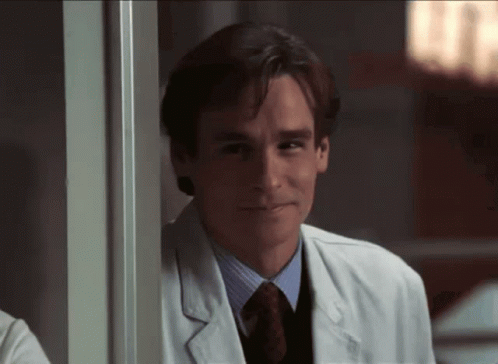 A Man In A Lab Coat And Tie Smiles While Leaning On A Wall GIF
