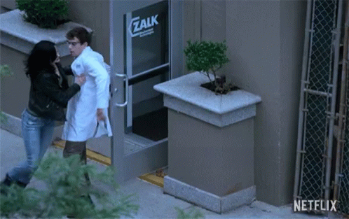 A Man In A Lab Coat Is Being Pushed By A Woman In Front Of A Zalk Building GIF