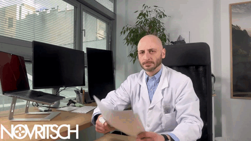 A Man In A Lab Coat Is Sitting At A Desk With The Word Novritsch On The Bottom Right GIF