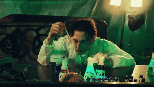A Man In A Lab Coat Is Sitting At A Table With A Bottle Of Vega On It GIF