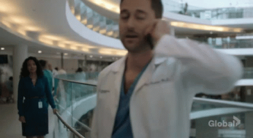 A Man In A Lab Coat Is Talking On A Cell Phone While Walking Down A Hospital Hallway . GIF