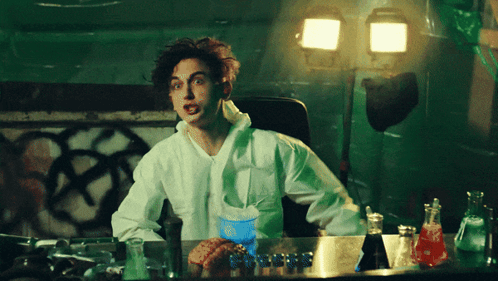 A Man In A Lab Coat Sits At A Desk With A Blue Cup That Says ' Coca Cola ' On It GIF