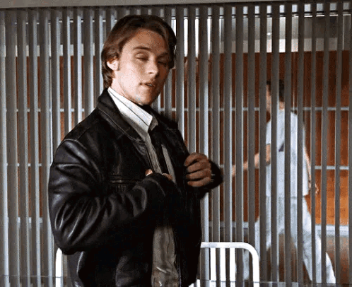 A Man In A Leather Jacket And Tie Is Standing In Front Of A Prison Cell . GIF