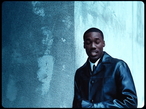 A Man In A Leather Jacket And Tie Stands In Front Of A Blue Wall GIF