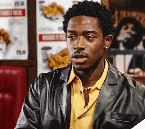 A Man In A Leather Jacket And Yellow Shirt Is Sitting In A Diner GIF