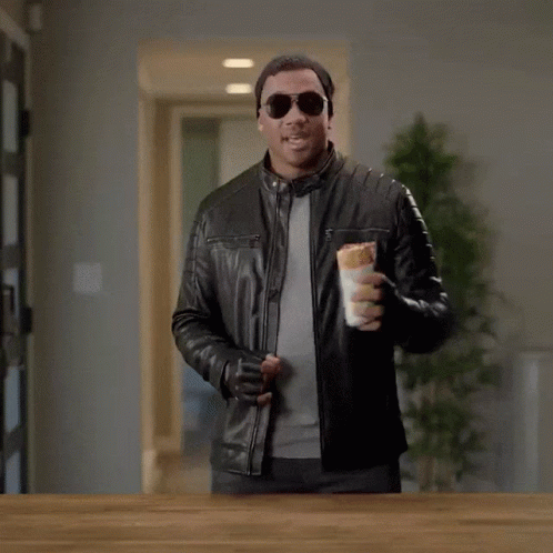 A Man In A Leather Jacket Holding A Sandwich With The Words " Here Comes The Airplane " On The Bottom GIF