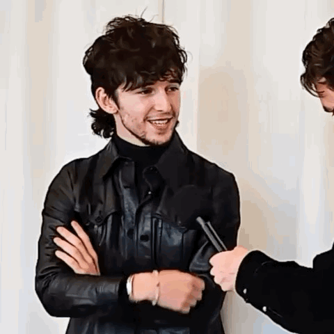 A Man In A Leather Jacket Is Being Interviewed By A Microphone While Standing Next To Another Man . GIF