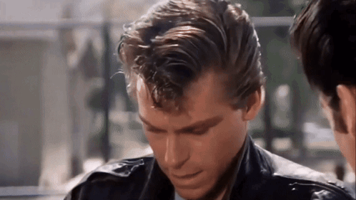 A Man In A Leather Jacket Is Looking Down At Something While Another Man Looks On . GIF