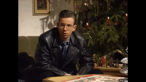 A Man In A Leather Jacket Is Sitting At A Table In Front Of A Christmas Tree . GIF