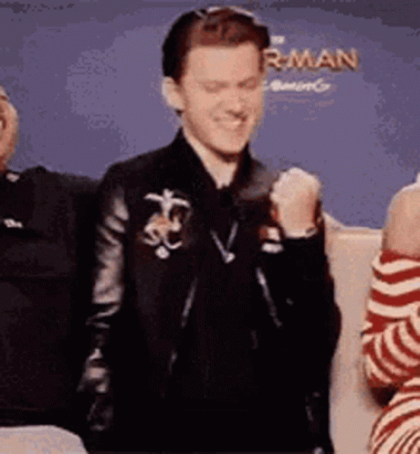 A Man In A Leather Jacket Is Sitting Next To A Woman In A Striped Shirt And Making A Fist . GIF