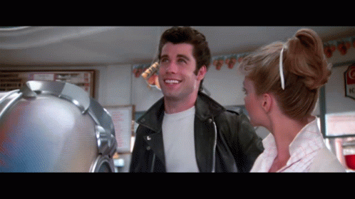A Man In A Leather Jacket Is Smiling Next To A Woman In A White Dress GIF