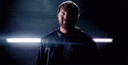 A Man In A Leather Jacket Is Standing In A Dark Room With A Light Shining On Him . GIF