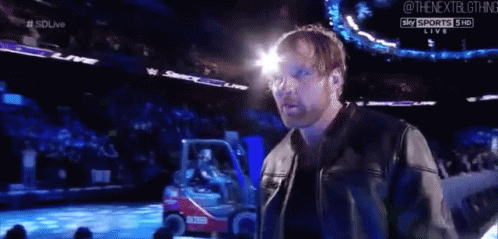 A Man In A Leather Jacket Is Standing In Front Of A Crowd At A Wrestling Event . GIF