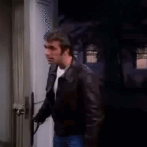 A Man In A Leather Jacket Is Standing In Front Of A Door And Holding A Gun . GIF