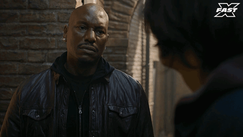 A Man In A Leather Jacket Is Talking To Another Man In Front Of A Brick Wall With X Fast Written On It GIF