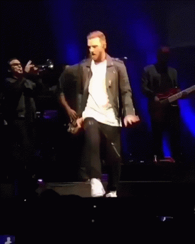A Man In A Leather Jacket Is Walking On A Stage With A Woman Behind Him GIF