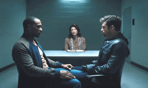 A Man In A Leather Jacket Sits In A Chair Talking To Two Other Men GIF