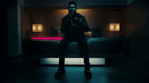 A Man In A Leather Jacket Sits On A Bed GIF