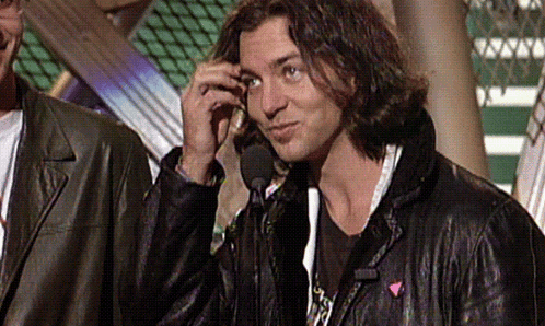 a man in a leather jacket talks into a microphone while another man stands behind him