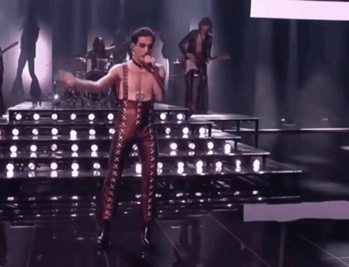 A Man In A Leather Outfit Is Singing Into A Microphone On Stage . GIF