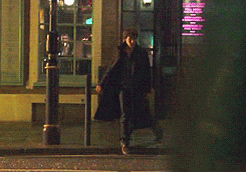 A Man In A Long Black Coat Is Walking Down A Street At Night GIF